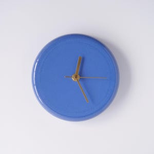 May include: A round, blue wall clock with gold-colored hands. The clock face is a solid, vibrant blue. The hands are thin and elegant, with a simple design. The clock is centered on a white background.