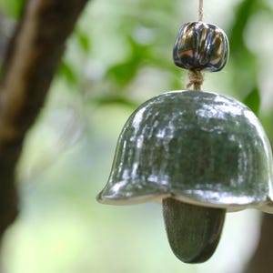 Puede incluir: Una campana de viento de cerámica hecha a mano con un cuerpo verde en forma de campana y una pieza superior más pequeña de color oscuro. La campana cuelga de un cordón de hilo natural, con un fondo verde borroso.