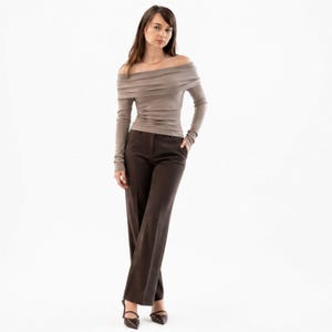 Pu&ograve; includere: Una donna indossa un top color taupe a maniche lunghe con spalle scoperte e pantaloni marrone scuro. I pantaloni sono a gamba larga. Indossa tacchi neri a punta. Lo sfondo &egrave; bianco.