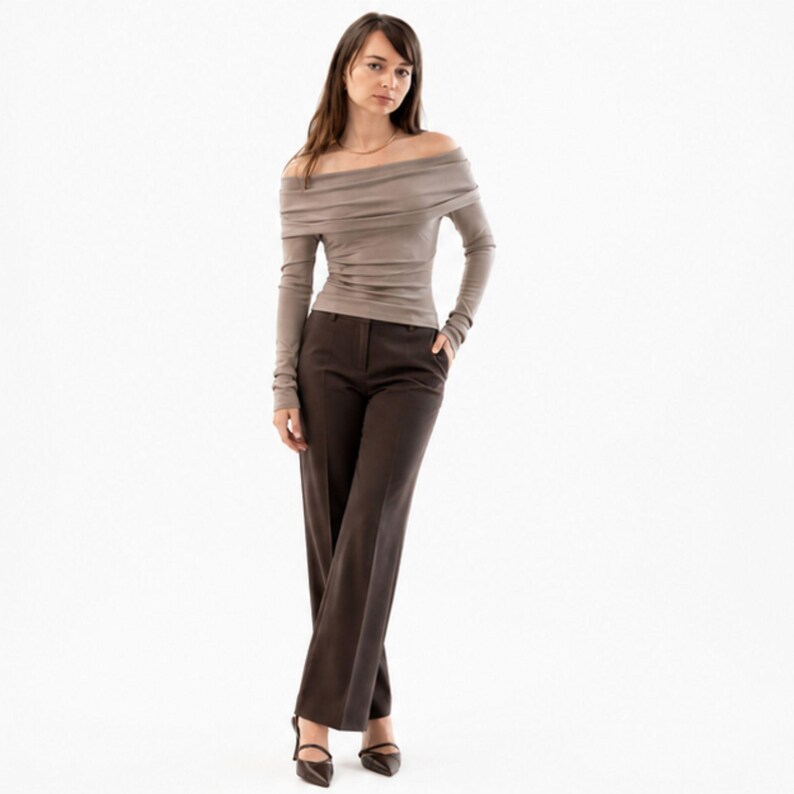 Pu&ograve; includere: Una donna indossa un top color taupe a maniche lunghe con spalle scoperte e pantaloni marrone scuro. I pantaloni sono a gamba larga. Indossa tacchi neri a punta. Lo sfondo &egrave; bianco.