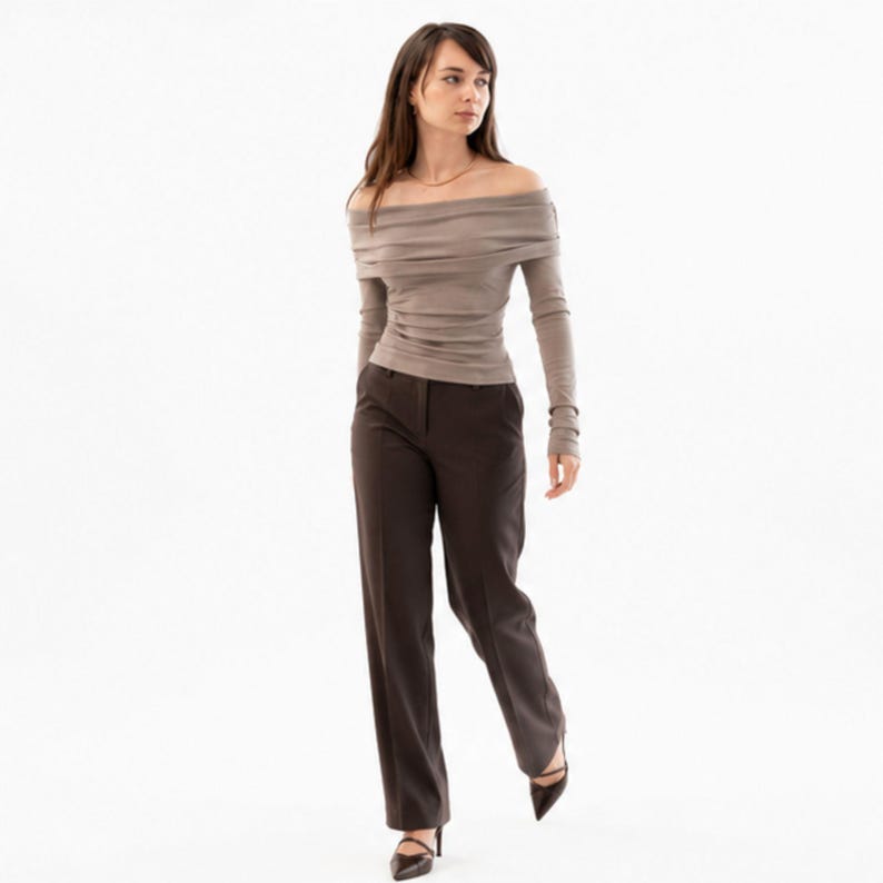 Pu&ograve; includere: Una donna indossa un top color talpa a maniche lunghe con spalle scoperte e pantaloni marrone scuro. I pantaloni sono a gamba dritta. Indossa tacchi neri a punta. Lo sfondo &egrave; bianco.
