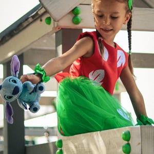 Peut inclure: Une enfant porte un costume rouge et vert, tenant un jouet en peluche bleu. Le costume comprend un haut rouge avec des détails blancs et une jupe en tulle verte. Une couronne de fleurs et des bracelets complètent la tenue. L'enfant sourit.