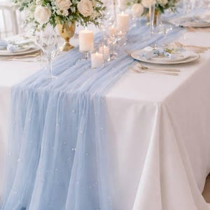 Könnte beinhalten: Ein festlich gedeckter Tisch mit weißer Tischdecke und einem hellblauen Tüllläufer, der mit kleinen, reflektierenden Akzenten besetzt ist. Blumengestecke in Goldvasen und Kerzen sorgen für ein elegantes Ambiente. Weingläser und Gedecke vervollständigen das Bild.