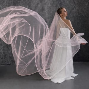May include: A long, flowing pale pink tulle wedding veil with a finished edge, worn with a strapless white floor-length gown. The sheer, lightweight fabric of the veil extends outward, creating a voluminous, airy effect behind the dress.