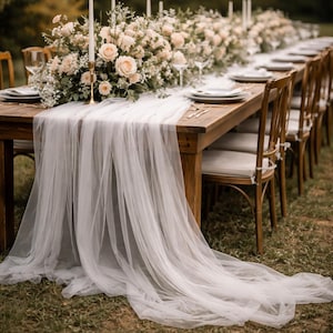Puede incluir: Una larga mesa de madera preparada para un evento formal, adornada con un camino de mesa de gasa blanca y centros de mesa florales. La mesa está puesta con platos, cubiertos y copas de vino, con sillas de madera dispuestas a ambos lados. Altas velas blancas añaden al ambiente elegante.