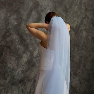 May include: A pale blue bridal veil flowing down the back of a white wedding dress. The veil is made of a sheer, delicate material. The dress has a fitted bodice and a flowing skirt. The background is a textured grey wall.