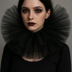 May include: A woman wearing a black, layered tulle collar. She has dark hair, dark eye makeup, and dark lipstick. The collar is the focal point, creating a dramatic, gothic aesthetic. The woman is wearing a black long-sleeved top.