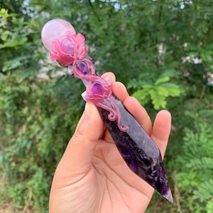 May include: A purple amethyst crystal wand with a pink crystal sphere at the top. The wand has pink, swirling, decorative accents. The wand is held in a hand against a background of green foliage.