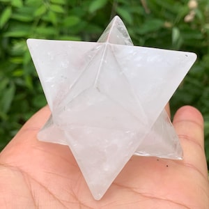 Op de afbeelding: Een heldere Merkaba-ster van kwartskristal. De geometrische ster wordt in een hand gehouden, met een groene, bladerrijke achtergrond. De Merkaba is een symbool van spirituele transformatie en wordt vaak gebruikt bij meditatie.