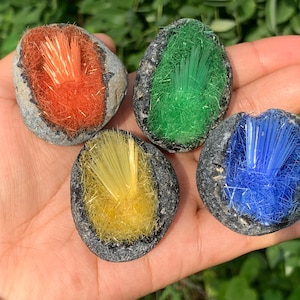 Può includere: Sono esposti quattro campioni minerali colorati. Ogni pietra ha un esterno grigio scuro e un interno vibrante arancione, verde, giallo e blu, con una consistenza a spillo simile a cristalli. Le pietre sono tenute in una mano.