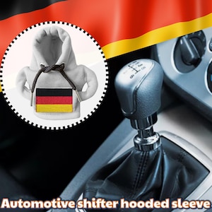 Puede incluir: Una funda para palanca de cambios de automóvil gris con forma de mini sudadera con capucha, con la bandera alemana. La funda tiene un cordón y está colocada contra el interior de un coche con una palanca de cambios.