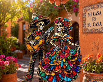 May include: A hanging acrylic ornament featuring two skeleton figures dressed in traditional attire. One figure wears a sombrero and plays a guitar, while the other wears a floral headpiece and a vibrant, multi-colored ruffled dress.