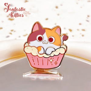 May include: A colorful enamel pin featuring a cartoon cat in a cupcake. The cat has red, white, and orange fur and is inside a pink cupcake with sprinkles. The pin has the text "Fantastic Kitties" in a cursive font.