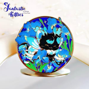 May include: A round enamel pin featuring a black and blue cartoon cat surrounded by blue iris flowers. The pin has a gold border and is set against a white background. The text "Fantastic Kitties" is at the top.