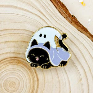 May include: A black cat enamel pin wearing a light blue ghost costume with a gold border. The cat has a smiling face with a pink tongue. The ghost costume has two black eye holes.