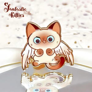 May include: A gold-colored enamel pin featuring a cartoon cat with angel wings. The cat has a cream-colored body, brown ears, and blue eyes. The pin has the text "Fantastic Kitties" in a cursive font.