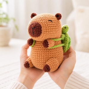 May include: A tan crochet capybara plush toy with a dark brown nose and feet, featuring a small green crochet turtle backpack attached to its back with light green straps. The capybara has embroidered eyes and a mouth, with a soft yarn texture.