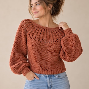 Puede incluir: Un suéter de ganchillo color óxido de manga larga con un canesú circular ancho y acanalado con detalles decorativos de ojales. La prenda tiene mangas globo voluminosas, cuello redondo y dobladillo acanalado, combinado con jeans azul claro.
