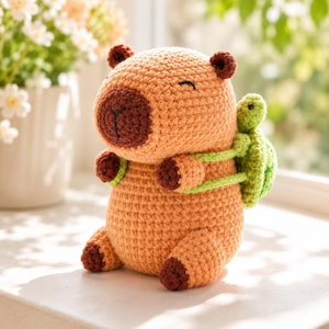 Puede incluir: Un juguete de capibara tejido a ganchillo en hilo naranja claro con hocico, orejas y patas de color marrón oscuro. El capibara tiene una expresión sonriente y lleva una pequeña tortuga tejida en verde claro a la espalda con correas verdes.