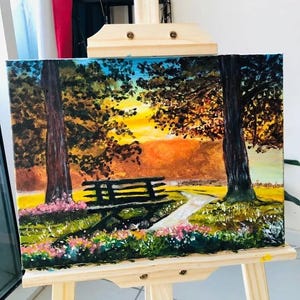 Puede incluir: Pintura al óleo sobre lienzo que representa una escena de parque con un banco, árboles y un camino. La obra presenta una vibrante puesta de sol con tonos de naranja, amarillo y azul. Flores rosas bordean el camino.
