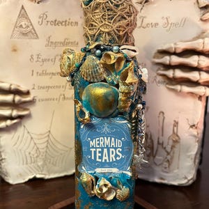 May include: A decorative bottle with a teal and gold finish, adorned with seashells and twine. The label reads "MERMAID TEARS" and "Est. 1886". The bottle is embellished with charms and beads, suggesting a mystical or apothecary theme.