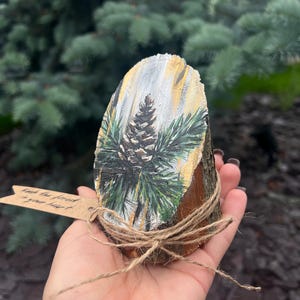 May include: A hand-painted wooden slice featuring a pine cone and evergreen branches. The artwork is on a natural wood slice, tied with twine, and includes a small tag with text. The color palette includes brown, green, white, and gold.