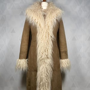 May include: A long, brown coat with a cream-colored, shaggy fur trim along the collar, front, cuffs, and hem. The coat is displayed on a mannequin, showcasing its full length and design. The coat appears to be made of suede or a similar material.