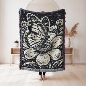 May include: A rectangular woven throw blanket featuring a high-contrast black and cream graphic design of a large butterfly resting on a flower. The blanket has a decorative fringed edge along the top and bottom, with a textured, knit-like appearance.