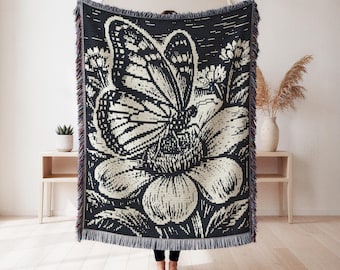 May include: A rectangular woven throw blanket featuring a high-contrast black and cream graphic design of a large butterfly resting on a flower. The blanket has a decorative fringed edge along the top and bottom, with a textured, knit-like appearance.