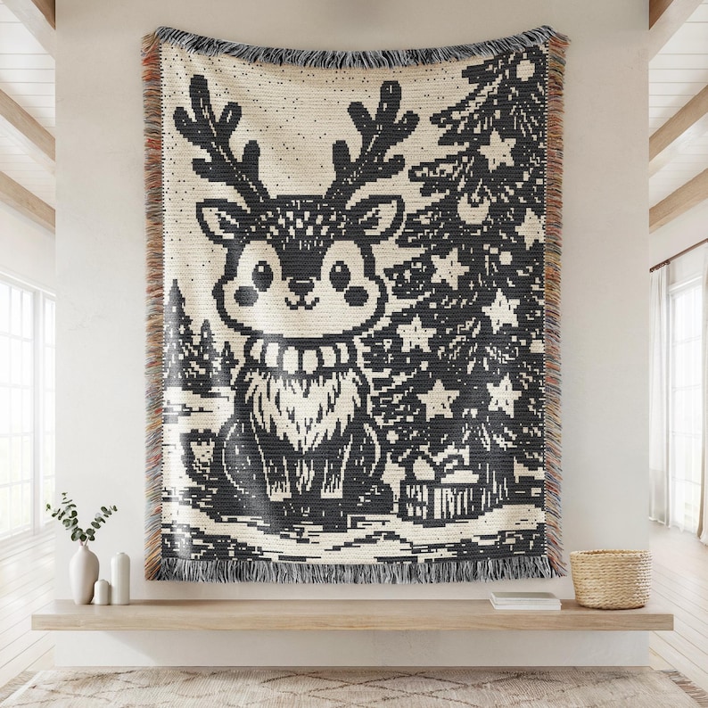 May include: A woven throw blanket featuring a black and cream graphic design of a reindeer with large antlers standing next to a decorated Christmas tree with star ornaments and a gift box, finished with a fringed edge on the top and bottom.