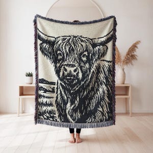 May include: A woven throw blanket featuring a high-contrast black and cream graphic of a Highland cow. The design displays the cow's shaggy fur, long horns, and facial features in a detailed pattern. The blanket has a fringed edge on all sides.