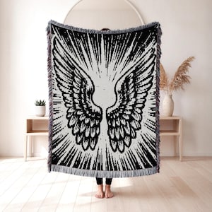 Puede incluir: Una manta tejida rectangular con un diseño gráfico en blanco y negro de alto contraste que muestra grandes alas de ángel detalladas en el centro. Las alas están rodeadas por líneas radiantes y los bordes tienen un acabado con flecos.