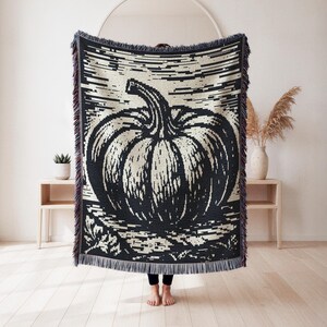 May include: A rectangular woven throw blanket featuring a large, high-contrast black and cream pumpkin graphic. The design includes horizontal line detailing in the background and a decorative fringe along the top and bottom edges of the textile.