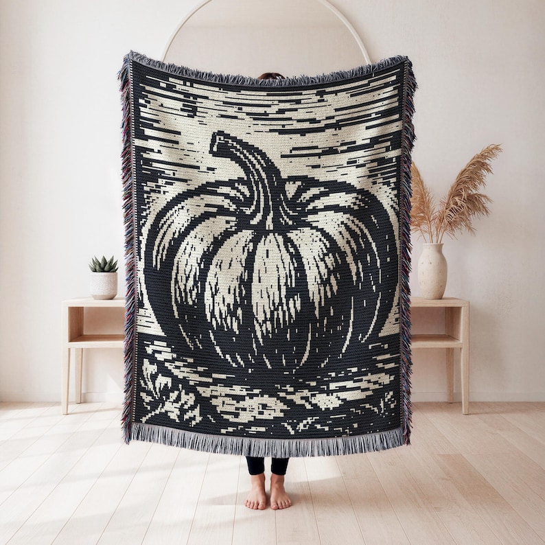 May include: A rectangular woven throw blanket featuring a large, high-contrast black and cream pumpkin graphic. The design includes horizontal line detailing in the background and a decorative fringe along the top and bottom edges of the textile.