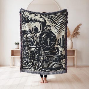 May include: A woven throw blanket featuring a high-contrast black and white graphic of a steam locomotive train. The design includes detailed wheels, a smokestack with billowing smoke, and a decorative fringed edge along the top and bottom.
