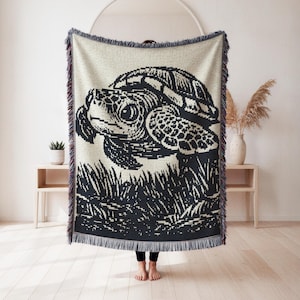 May include: A woven throw blanket featuring a detailed black and white illustration of a sea turtle in a grassy area. The blanket has a cream-colored background with a fringed edge in shades of blue, purple, and red. The turtle's shell and head are intricately designed.