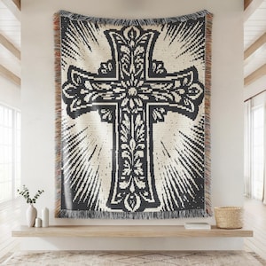 May include: A woven throw blanket featuring a large, dark charcoal cross centered on a cream background. The cross is decorated with intricate floral and leaf patterns, surrounded by radiating sunburst lines. The edges have a multi-colored fringe.