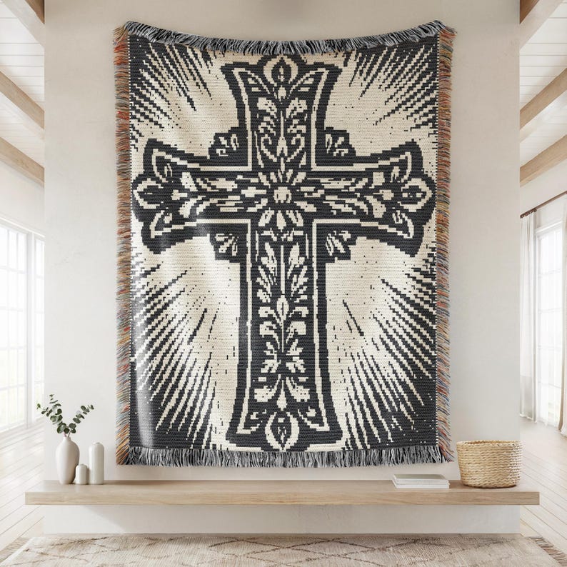 May include: A woven throw blanket featuring a large, dark charcoal cross centered on a cream background. The cross is decorated with intricate floral and leaf patterns, surrounded by radiating sunburst lines. The edges have a multi-colored fringe.