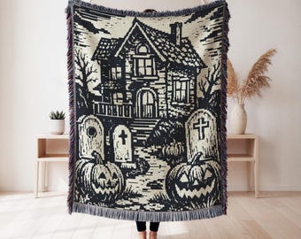 May include: A woven throw blanket featuring a black and cream graphic design of a haunted house, tombstones, and jack-o'-lanterns. The rectangular blanket has a fringed edge and displays a detailed scene of a spooky dwelling with bare trees.