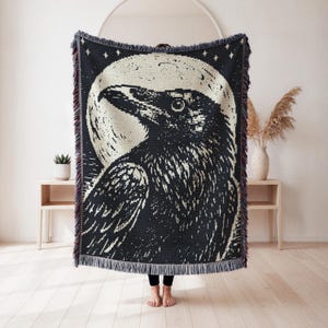 May include: A woven throw blanket featuring a large black raven centred against a full moon background. The design includes small star motifs at the top and a decorative fringed edge along the bottom. The colour palette consists of black and cream.