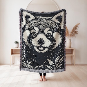 May include: A woven throw blanket featuring a high-contrast black and cream graphic of a red panda face. The rectangular textile has a textured, knit-like appearance with decorative fringe along the top and bottom edges, showcasing a detailed animal portrait.