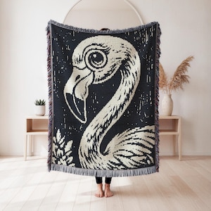 May include: A woven throw blanket featuring a large, cream-colored flamingo graphic set against a solid black background. The blanket has a textured, pixelated appearance with decorative fringe along the top and bottom edges in dark tones.