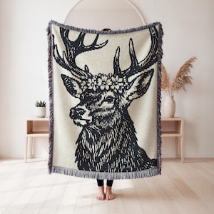 May include: A woven throw blanket featuring a large black and white graphic of a deer head with a floral crown between its antlers. The rectangular blanket has a textured knit appearance and is finished with decorative fringe along the edges.