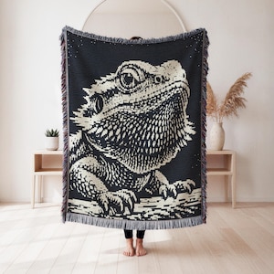 May include: A woven throw blanket featuring a high-contrast black and cream graphic of a bearded dragon lizard. The rectangular textile has a textured surface with a fringed edge along the top and bottom, displaying a detailed reptile portrait.