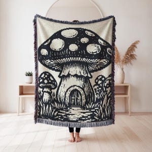 Peut inclure: Un plaid tissé rectangulaire présentant un grand motif central de champignon noir et crème avec une petite porte arquée à la base. Le design comprend des champignons plus petits sur les côtés et un bord à franges décoratif sur les quatre côtés.