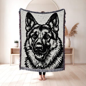 Puede incluir: Una manta tejida que presenta un retrato pixelado en blanco y negro de alto contraste de un perro pastor alemán. La manta tiene una superficie texturizada con bordes deshilachados en la parte superior e inferior, mostrando un rostro canino.