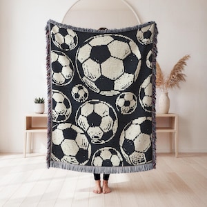 May include: A rectangular woven throw blanket featuring a repeating pattern of black and white soccer balls. The blanket has a dark background with multiple soccer ball graphics of varying sizes and includes decorative fringe along the top and bottom edges.
