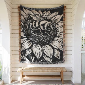 May include: A woven throw blanket featuring a large, high-contrast black and cream graphic of a bee resting on a sunflower. The rectangular textile has a textured knit pattern and is finished with decorative multi-colored fringe on all sides.