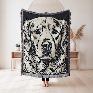 Op de afbeelding: Een geweven plaid met een groot, contrastrijk zwart en crèmekleurig grafisch portret van een golden retriever. De rechthoekige deken heeft een getextureerde gebreide constructie en is afgewerkt met decoratieve franjes langs de randen.