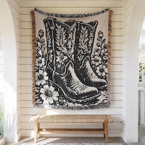 Puede incluir: Una manta tejida con un diseño gráfico en negro y crema de dos botas vaqueras rodeadas de motivos florales. El tejido rectangular tiene un borde con flecos y hilos multicolores en los lados izquierdo y derecho.